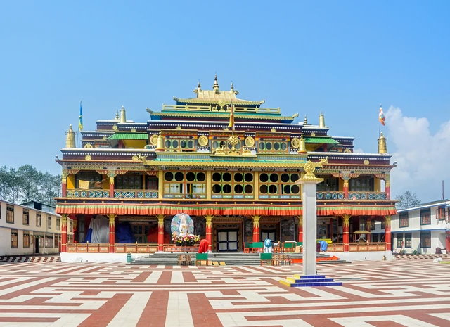 The painted main hall of Ralang Monastery with golden roofs and a tall pillar in front on a red and white paved courtyard under a blue sky