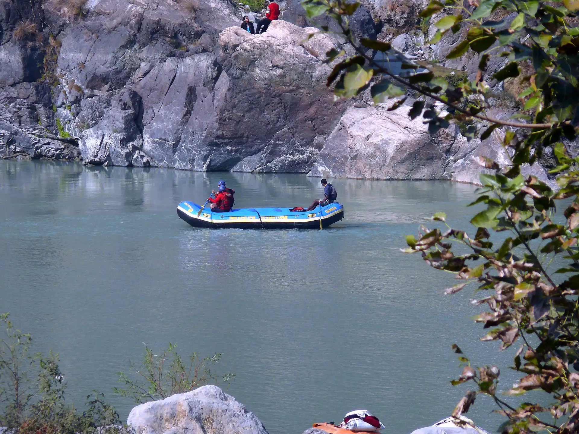 A small blue raft on the clear green Ganga between grey rocks and overhanging branches near Rishikesh