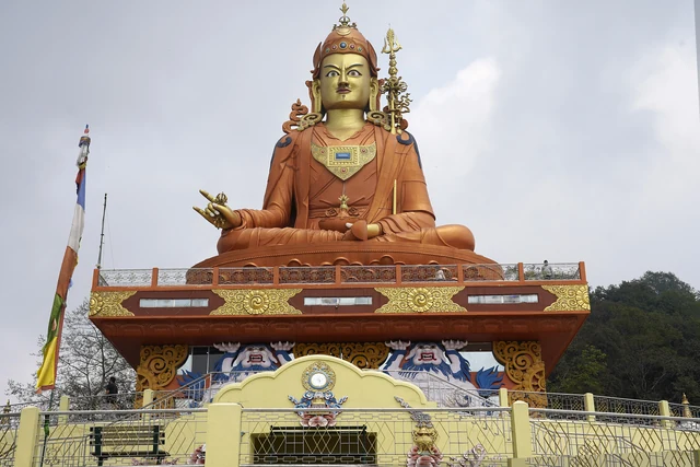 The seated golden statue of Guru Padmasambhava above a yellow and red painted base with a prayer flag pole beside it
