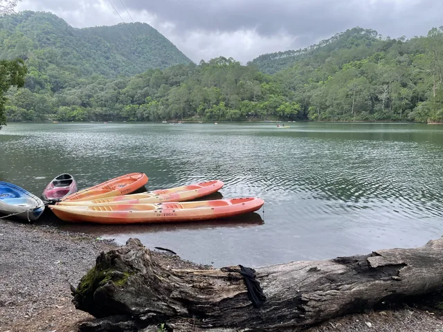 Brightly coloured kayaks on the stony shore of Sattal lake, with green forested hills and a cloudy sky behind