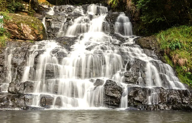 A broad, tiered waterfall spilling white over dark rock ledges edged with ferns and green moss at Elephant Falls