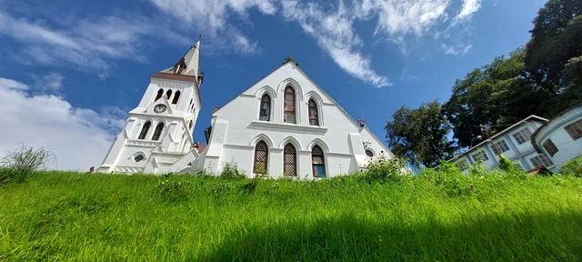 The white gabled front and clock tower of St Andrew's Church seen from a grassy bank under a blue sky with clouds