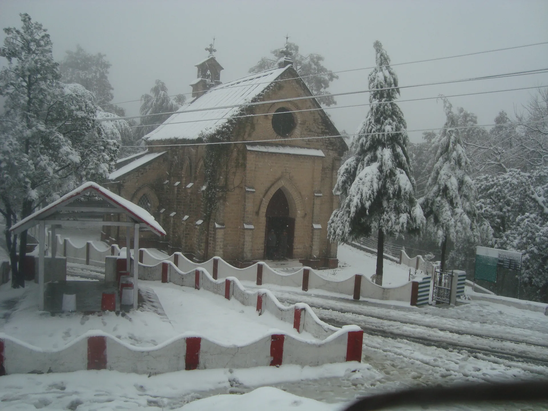 St Mary's Church and the deodars around it covered in fresh snow, with a snowy road and railings in front