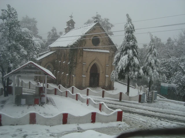 St Mary's Church and the deodars around it covered in fresh snow, with a snowy road and railings in front