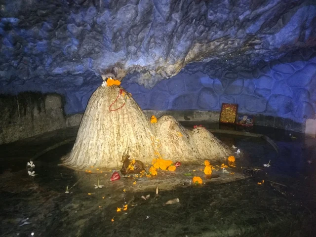 A cone of white-wrapped shivalinga with orange flowers inside a blue-lit cave at Tapkeshwar