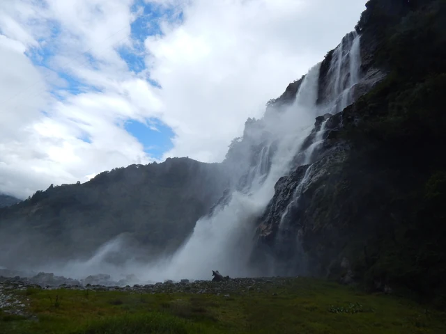Nuranang Falls pouring down a high green cliff in a wide white plume, with cloud and blue sky above