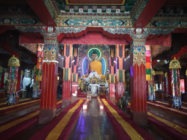 Inside the prayer hall of Tawang Monastery: red pillars, hanging banners and rows of seats leading to a golden image of the Buddha