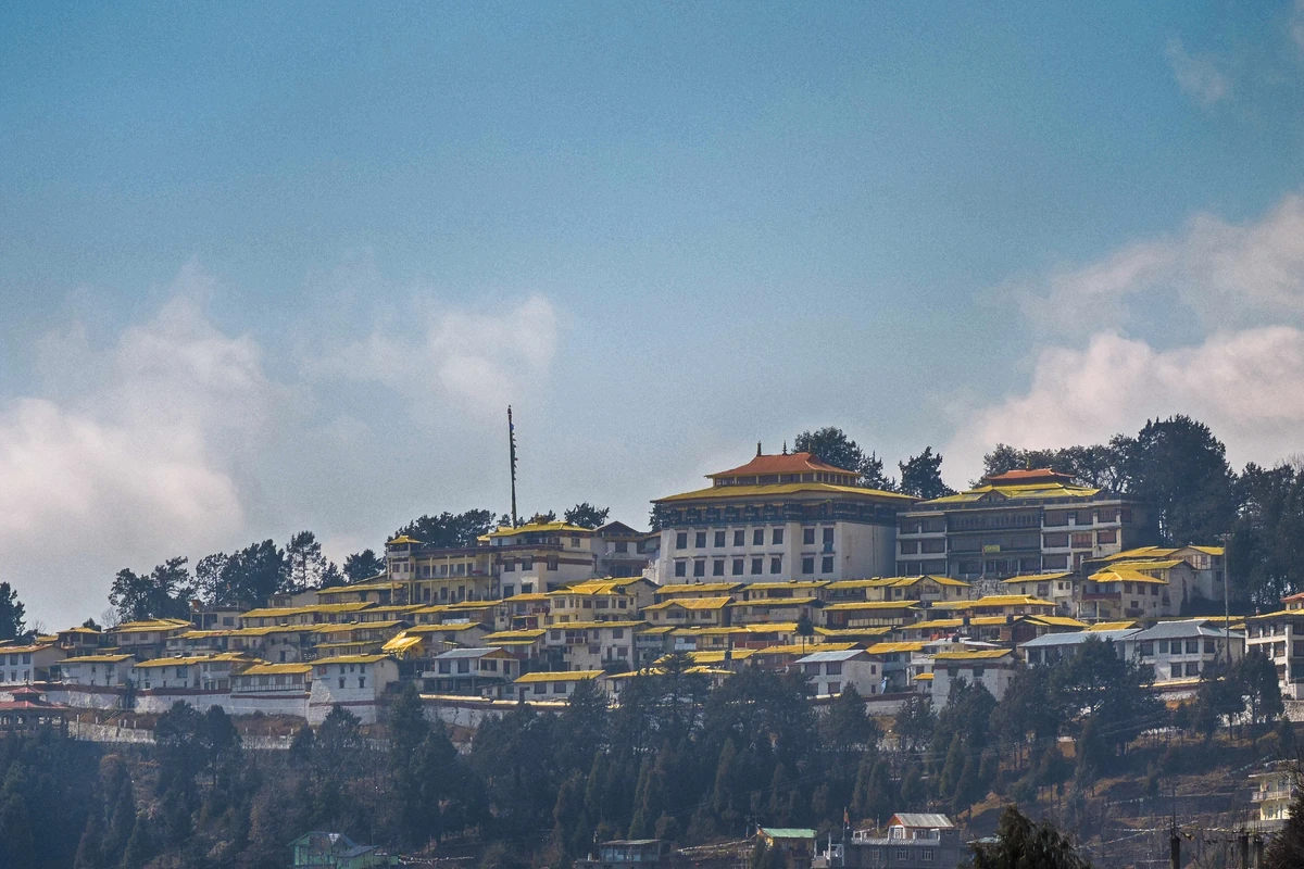 The white and yellow buildings of Tawang Monastery spread along a ridge above the town under a blue sky with clouds