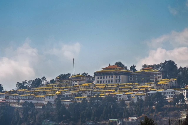 The white and yellow buildings of Tawang Monastery spread along a ridge above the town under a blue sky with clouds