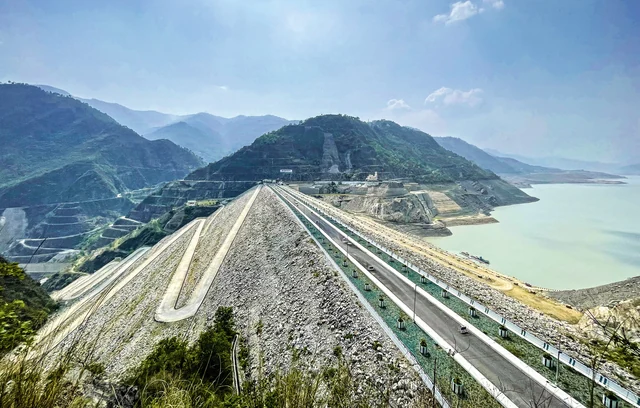 The long rock-and-earth crest of the Tehri Dam with its road and the reservoir behind, under a blue sky