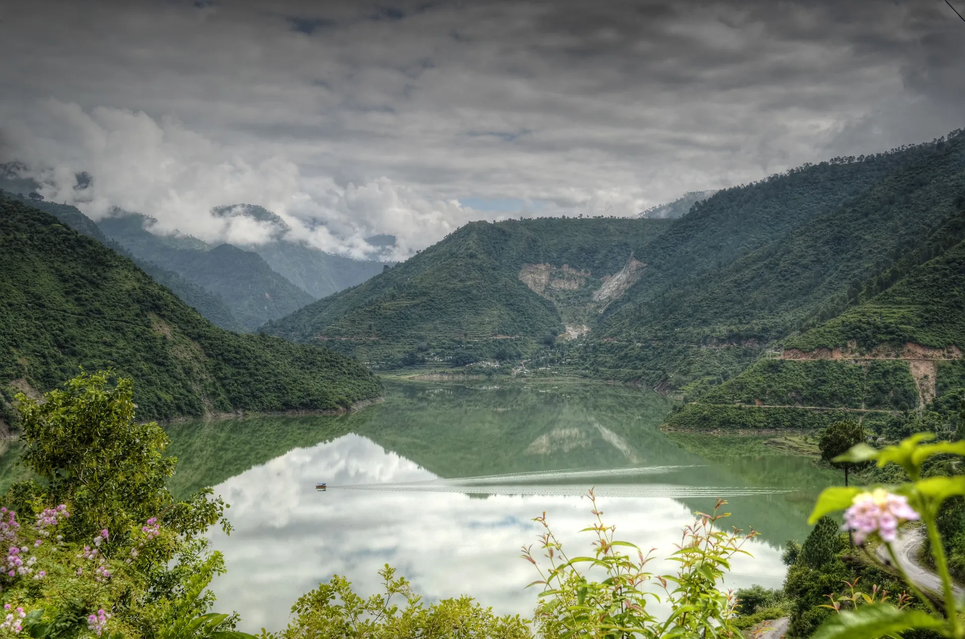 The still Tehri lake reflecting cloudy hills, framed by flowers and green slopes