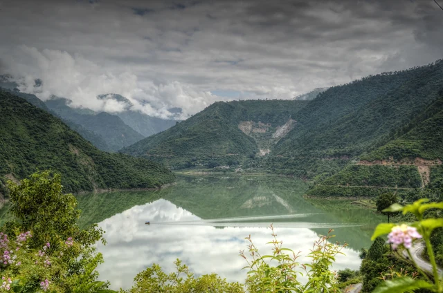 The still Tehri lake reflecting cloudy hills, framed by flowers and green slopes