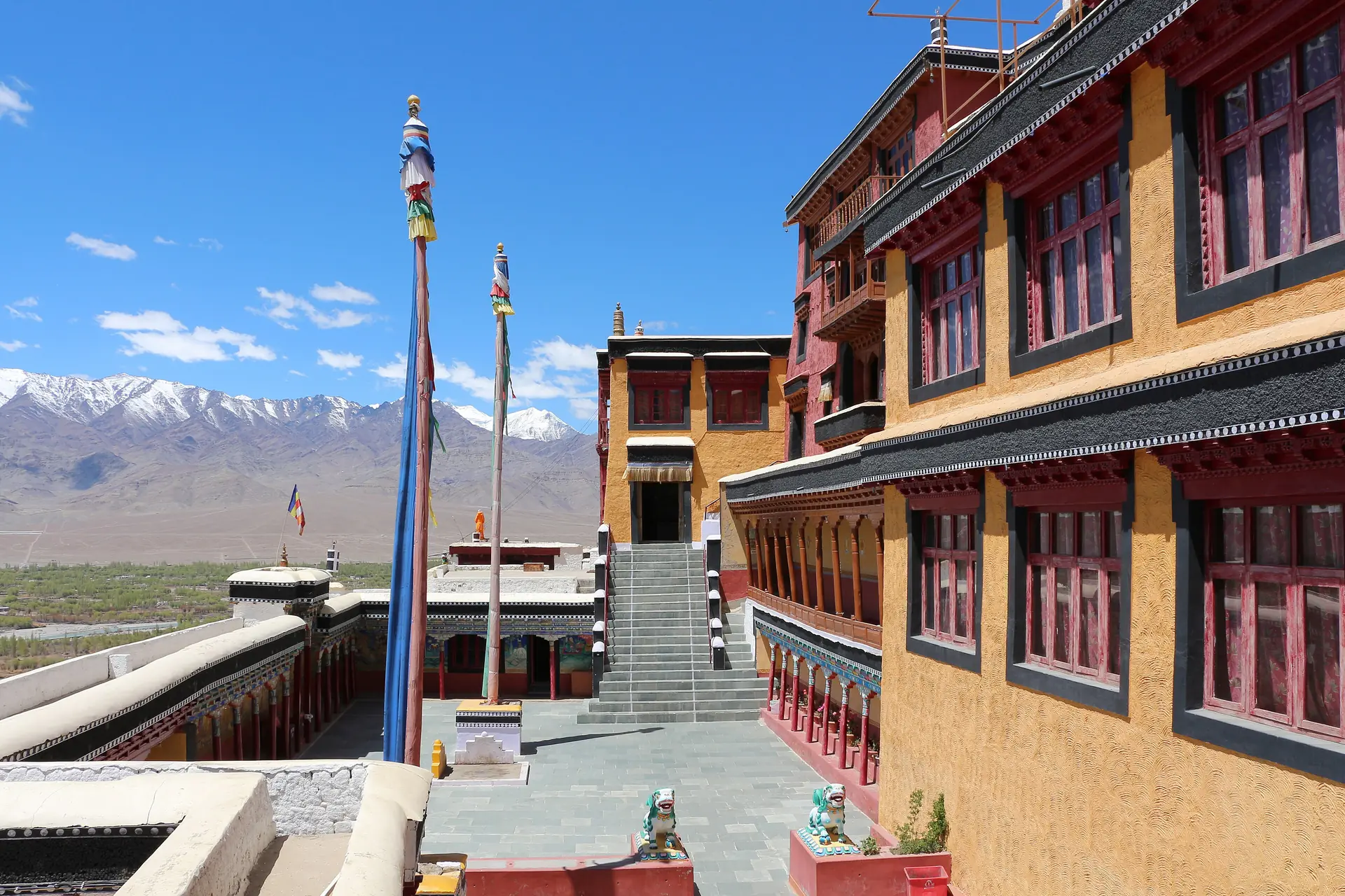 Red, yellow and white monastery buildings with flagpoles and a stairway, and snow peaks along the horizon