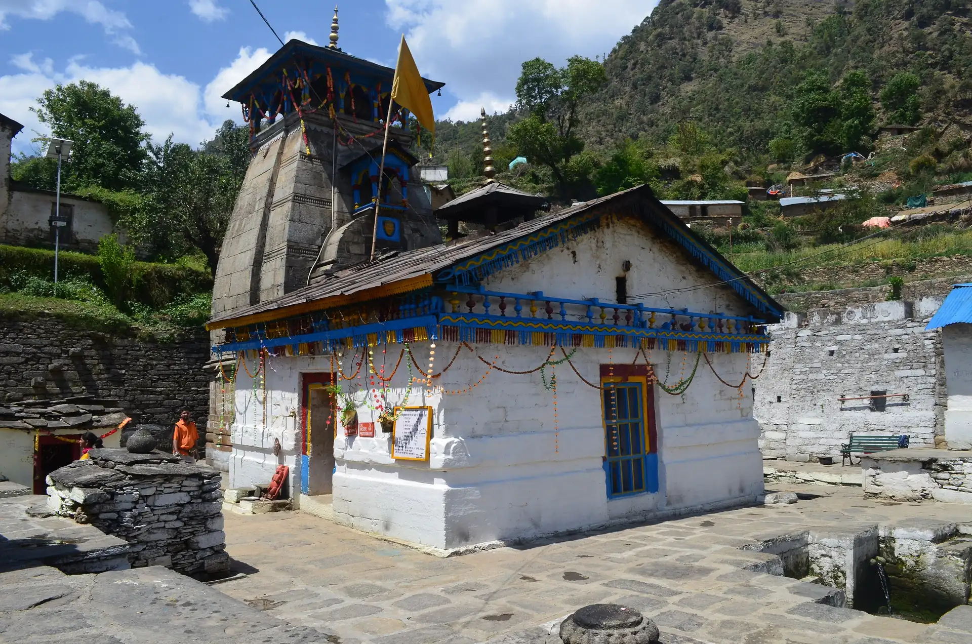 The small white and blue Triyuginarayan temple with a dark stone tower, its paved courtyard and a flag