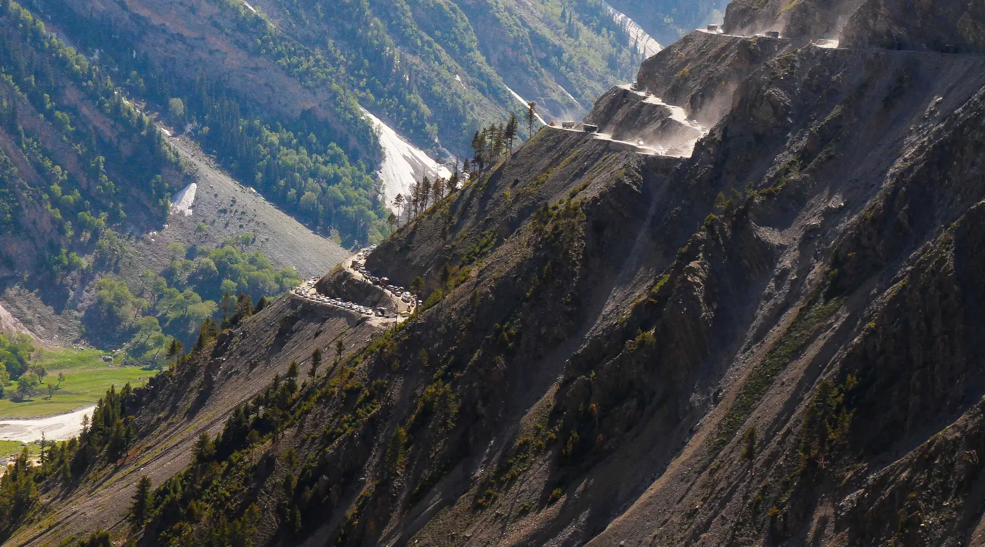 A narrow mountain road cut in hairpin switchbacks across a steep dry slope above a green valley at Zoji La