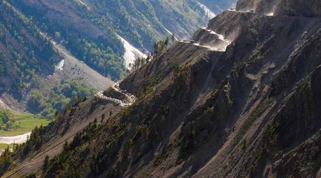 A narrow mountain road cut in hairpin switchbacks across a steep dry slope above a green valley at Zoji La