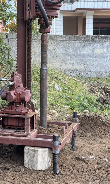 Close-up of the portable rig drilling: the drill rod going down through the frame, a screw jack on a block beside it