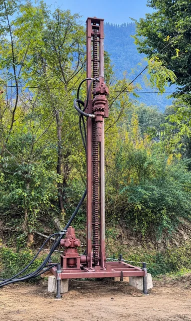 The portable rig standing on its steel frame: a tall chain-fed mast, the rotary head halfway up, hydraulic hoses, screw jacks on concrete blocks