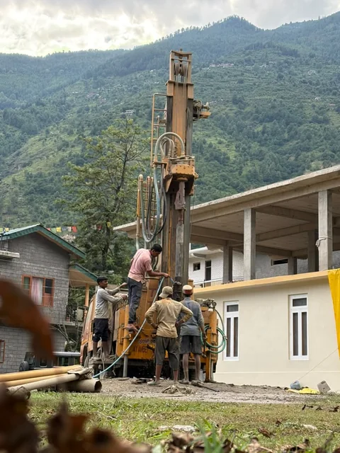 The rig's mast up beside a new building, the crew at the rods, terraced hills behind